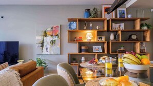 Un salon chaleureux avec un canapé, des fauteuils, une table basse sur laquelle sont disposés des éléments pour le petit-déjeuner, et une bibliothèque contenant divers objets décoratifs.
