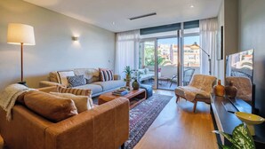 Un salon moderne avec un canapé, des fauteuils, une table basse et un téléviseur. Il y a un balcon avec vue sur les immeubles et une plante sur le rebord de la fenêtre.