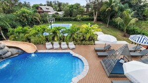 A swimming pool with a waterfall feature, surrounded by lounge chairs and umbrellas, set in a lush garden with tropical plants. - Glam Manor | 7,000 Ft Mansion with Waterslide | Sauna | Cold plunge (Sunrise)
