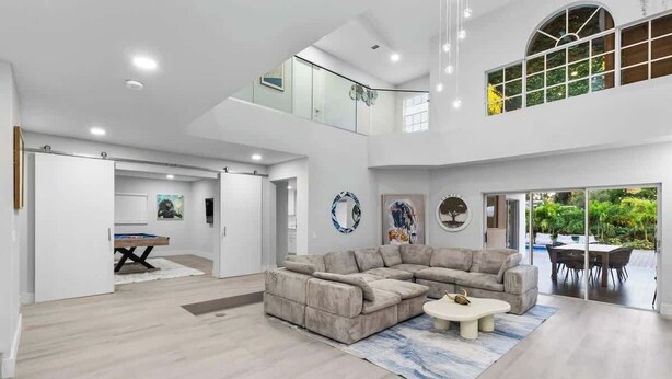 A spacious living area with a large L-shaped sectional sofa, a coffee table, and a dining table with chairs. There is a staircase leading to an upper level, and a view of a garden through large windows. - Glam Manor | 7,000 Ft Mansion with Waterslide | Sauna | Cold plunge (Sunrise)
