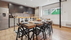 A modern kitchen with dark cabinets, a wooden dining table set for six, and a view of a green outdoor area through a window. - Suite 332 Brady – Stunning Duplex in Paris 10 (Paris)