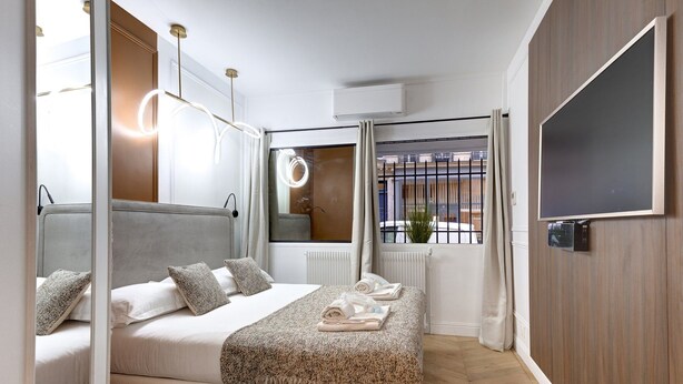 Een moderne slaapkamer met een bed, nachtlampjes, een spiegel, een raam met gordijnen, en een televisie die aan de muur is bevestigd.