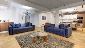 Een ruime woonkamer met twee banken, een glazen salontafel en een keuken op de achtergrond.
