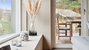 Ein modernes Wohnzimmer mit großem Fenster, einer Glasvase mit Pampasgras, einem dekorativen Steinglas und Blick auf eine Terrassenanlage.