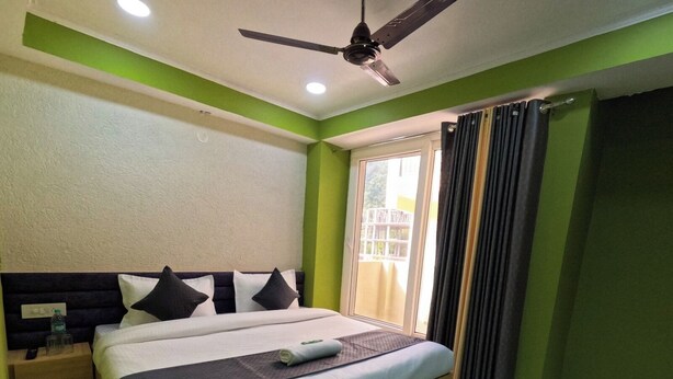 A bedroom with a bed, a nightstand, a ceiling fan, and a window with curtains. - Hotel Aastha Height (Rishikesh)