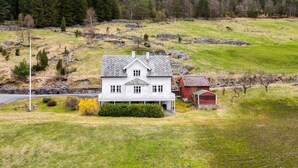 A white house with a porch, surrounded by a green landscape and a stone wall.