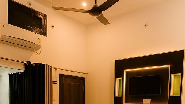 A hotel room with a bed, a bedside table, a TV mounted on the wall, an air conditioning unit, and a ceiling fan. - Ramam By Naavagat 700 Meter From Temple (Faizabad)