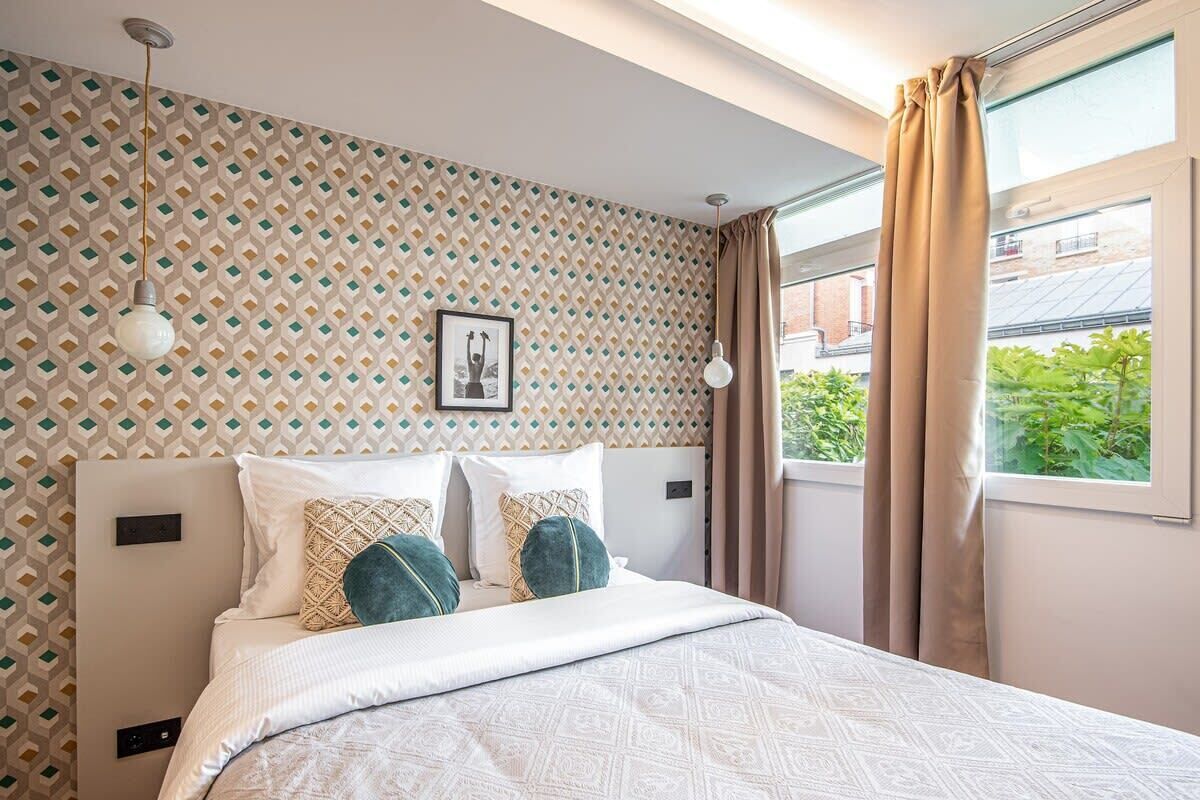 Ein Schlafzimmer mit gemustertem Wandpapier, einem Bett mit weißer Bettwäsche und dekorativen Kissen, einem gerahmten Bild an der Wand und einem Fenster mit Vorhängen.