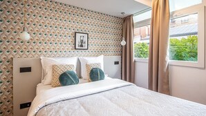 Ein Schlafzimmer mit gemustertem Wandpapier, einem Bett mit weißer Bettwäsche und dekorativen Kissen, einem gerahmten Bild an der Wand und einem Fenster mit Vorhängen.