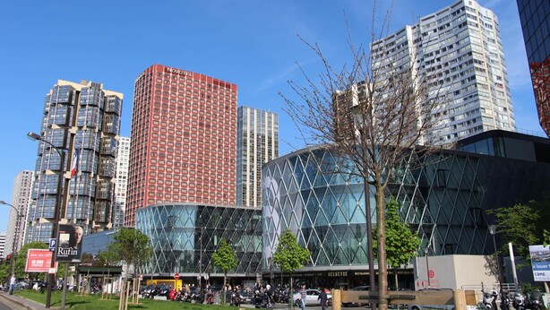 Eine moderne Stadtstraße mit Hochhäusern, darunter ein markantes rotes Gebäude und eine Glasstruktur mit geometrischem Muster.