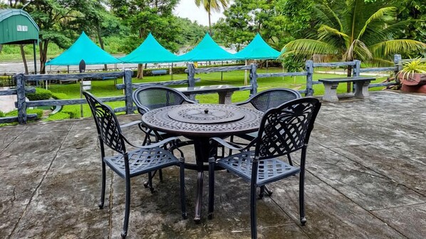 A round metal table with four chairs, surrounded by a stone patio with a wooden fence and green grass. - Sakthi River Resorts (Anaimalai)