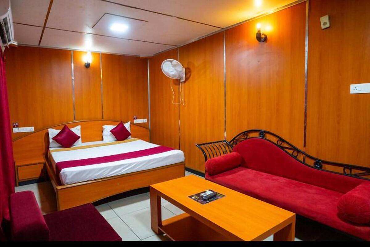 A hotel room with a bed, a red sofa, a wooden table, and a wall-mounted fan.