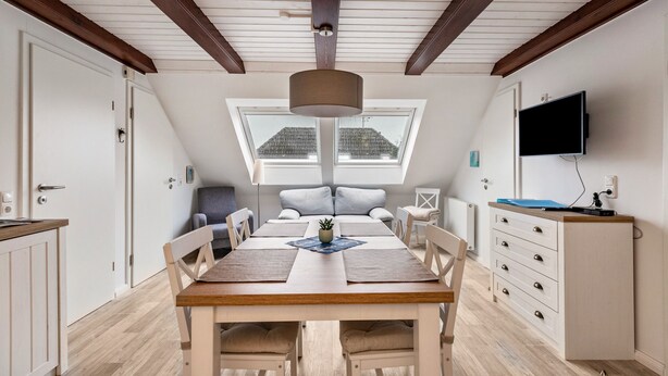 A dining area with a wooden table, chairs, a ceiling fan, a TV mounted on the wall, and a window with a view of a house.