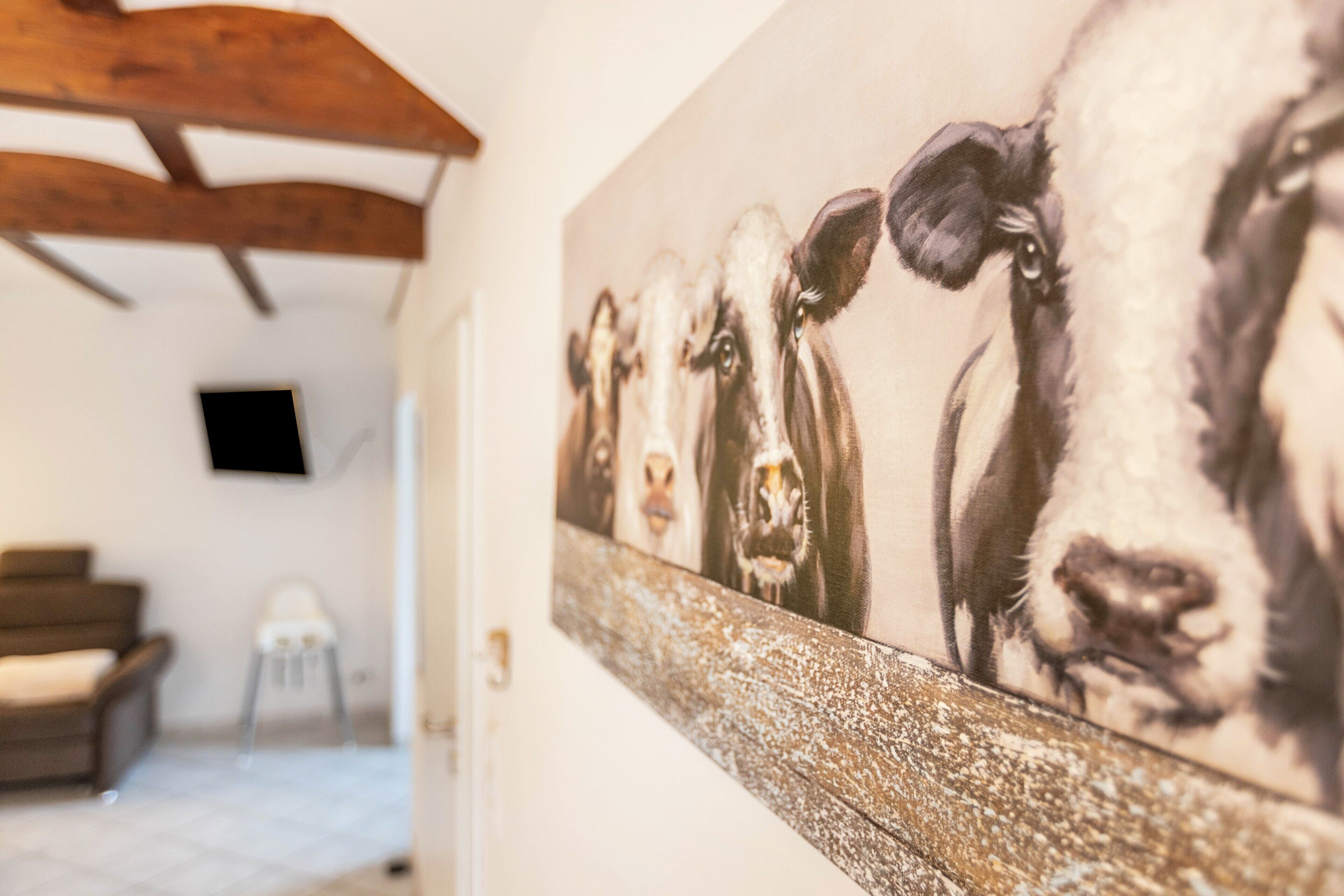 Corredor com uma obra de arte na parede retratando vacas, um vigamento de madeira no teto, uma cadeira e uma televisão.