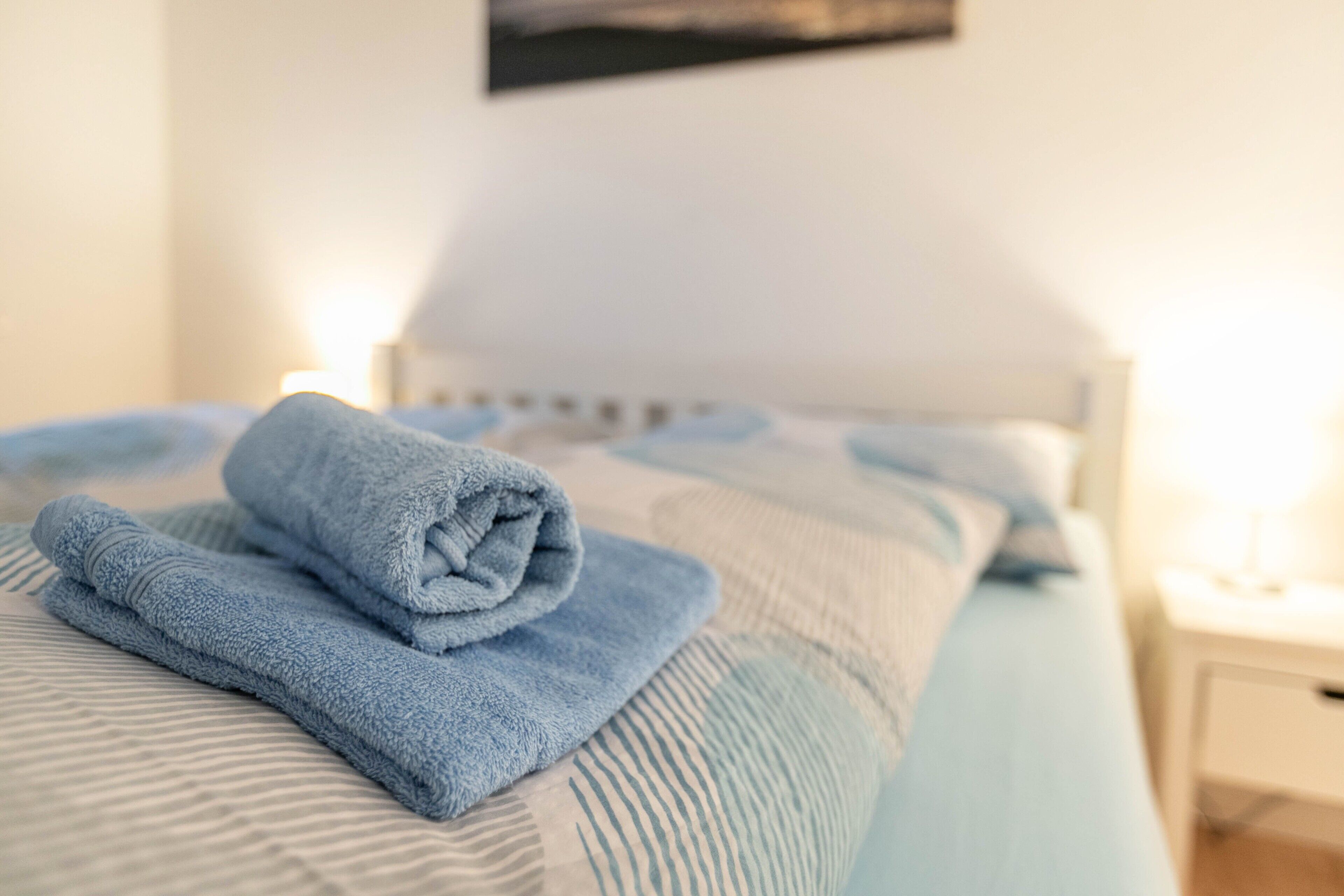 Ein ordentlich bezogenes Bett mit einem gefalteten blauen Handtuch, einem gestreiften Kopfkissen und einem weißen Kopfteil.