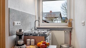 Een kleine keuken met een koffiezetapparaat, een schaal fruit, een spoelbak en een raam met uitzicht op een huis.