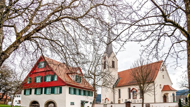 Een klassiek gebouw met een steile kap, een kerk met een hoge torenspits, en een kleiner gebouw met een rood dak.