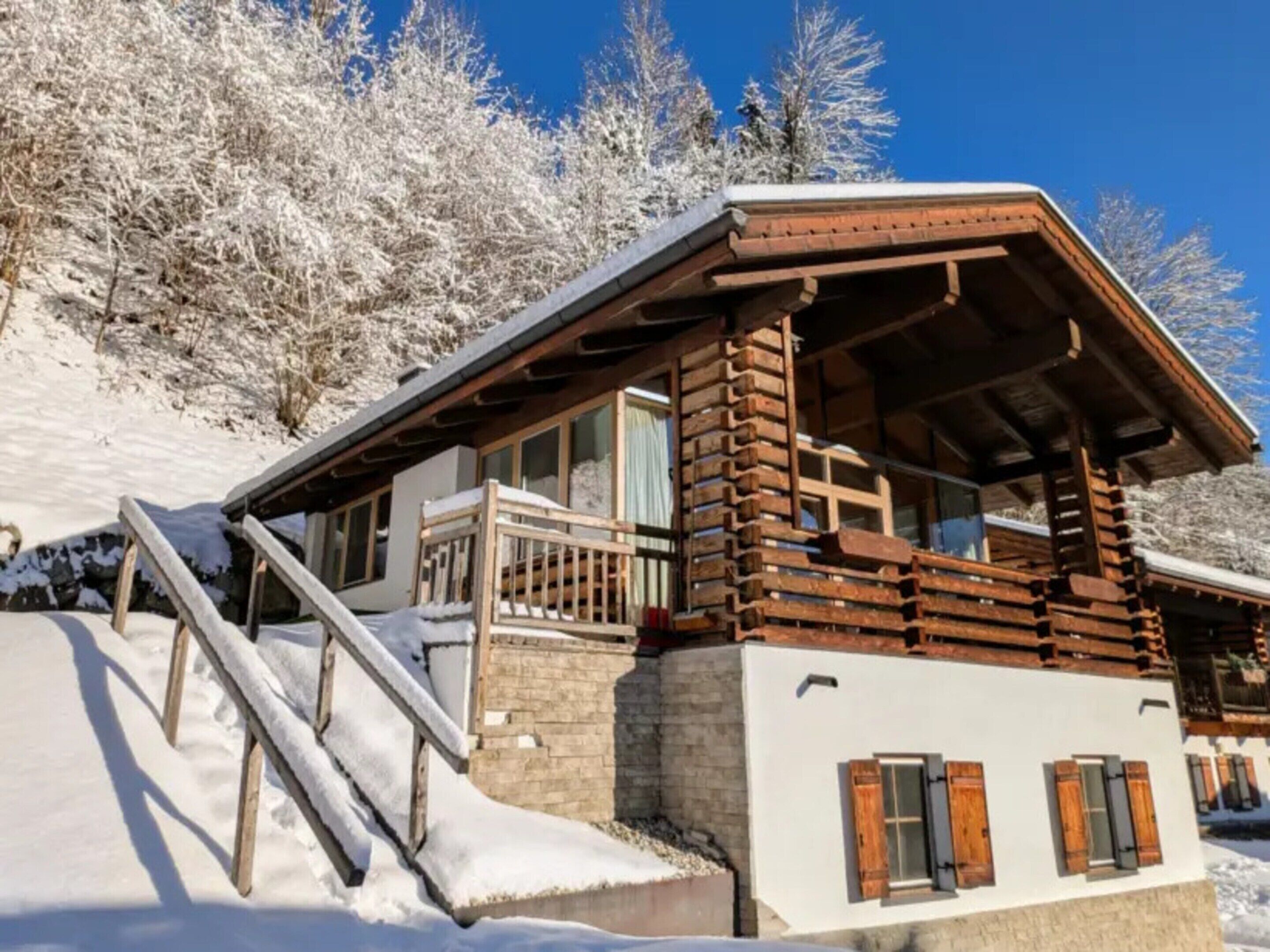 Una cabaña de madera con un balcón, una escalera que lleva a él, y árboles cubiertos de nieve al fondo.