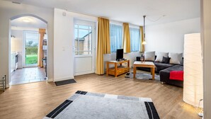 Un salón moderno con sofá, televisión y lámpara. El suelo es de madera y hay una puerta que da a una cocina.