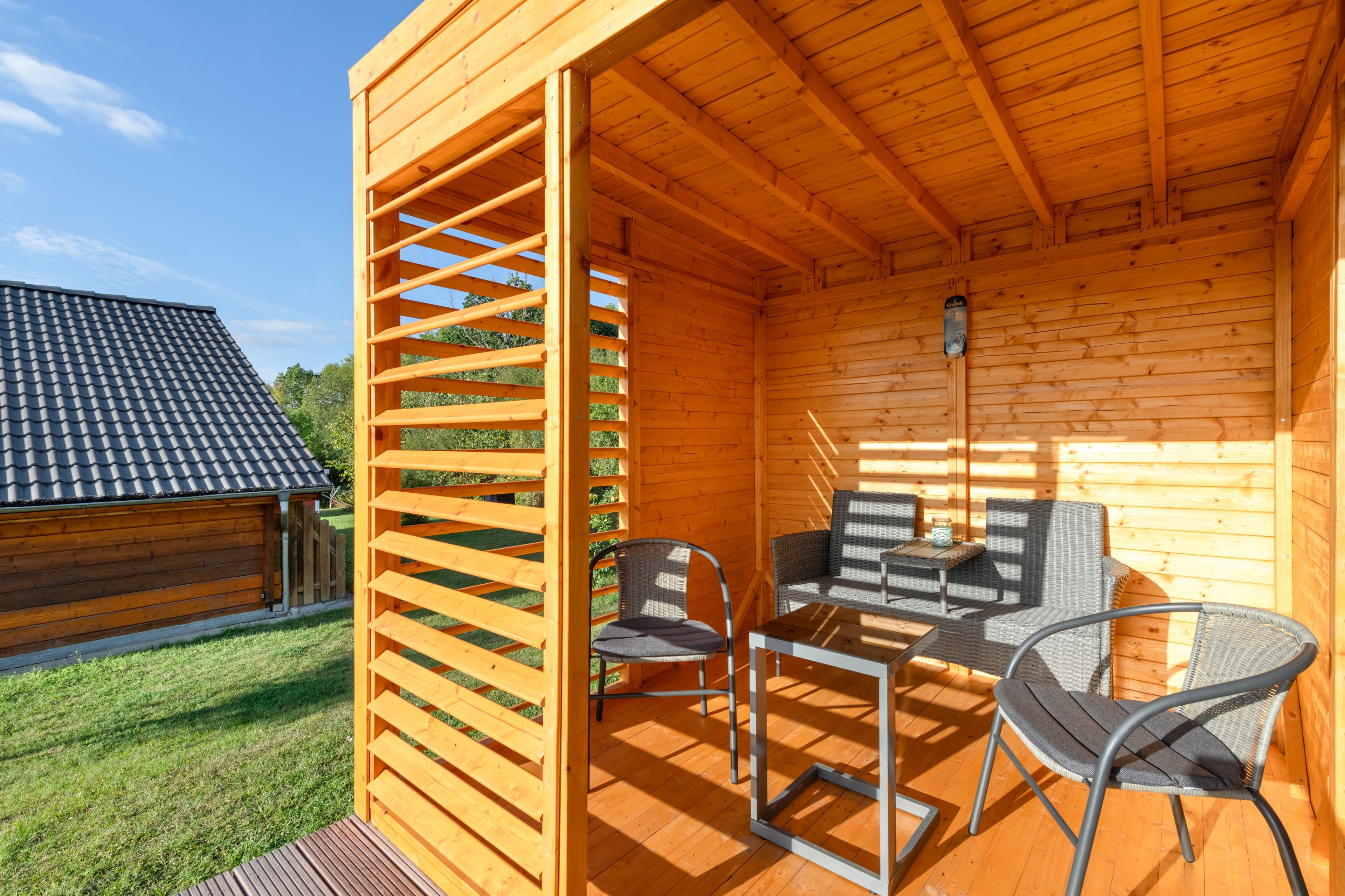 A wooden cabin with a patio area featuring a table and chairs, a wooden fence, and a view of a neighboring building.