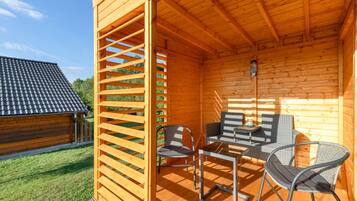 A wooden cabin with a patio area featuring a table and chairs, a wooden fence, and a view of a neighboring building.