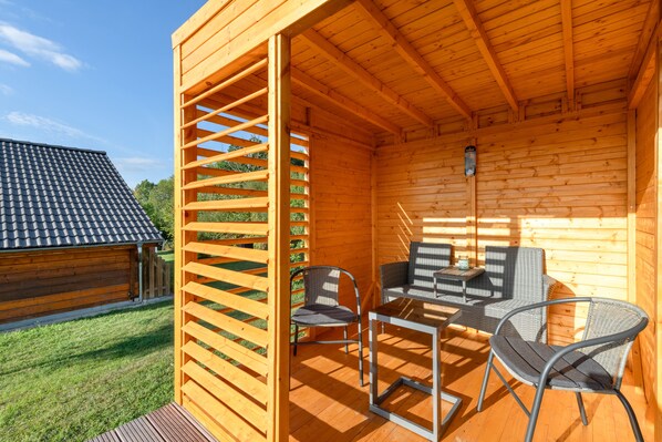 A wooden cabin with a patio area featuring a table and chairs, a wooden fence, and a view of a neighboring building.