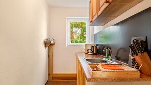 Cocina compacta con armarios de madera, ventana con vistas a árboles, un lavamanos y una encimera con tabla de cortar y zanahorias.
