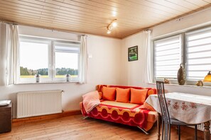 A room with a wooden floor, a white radiator, a sofa with orange cushions, a table with a white tablecloth, and a window with a view of greenery. - Apartment 'Möblierte Wohnung Ferienwohnung' with Shared Terrace, Shared Garden and Wi-Fi (Aicha vorm Wald)