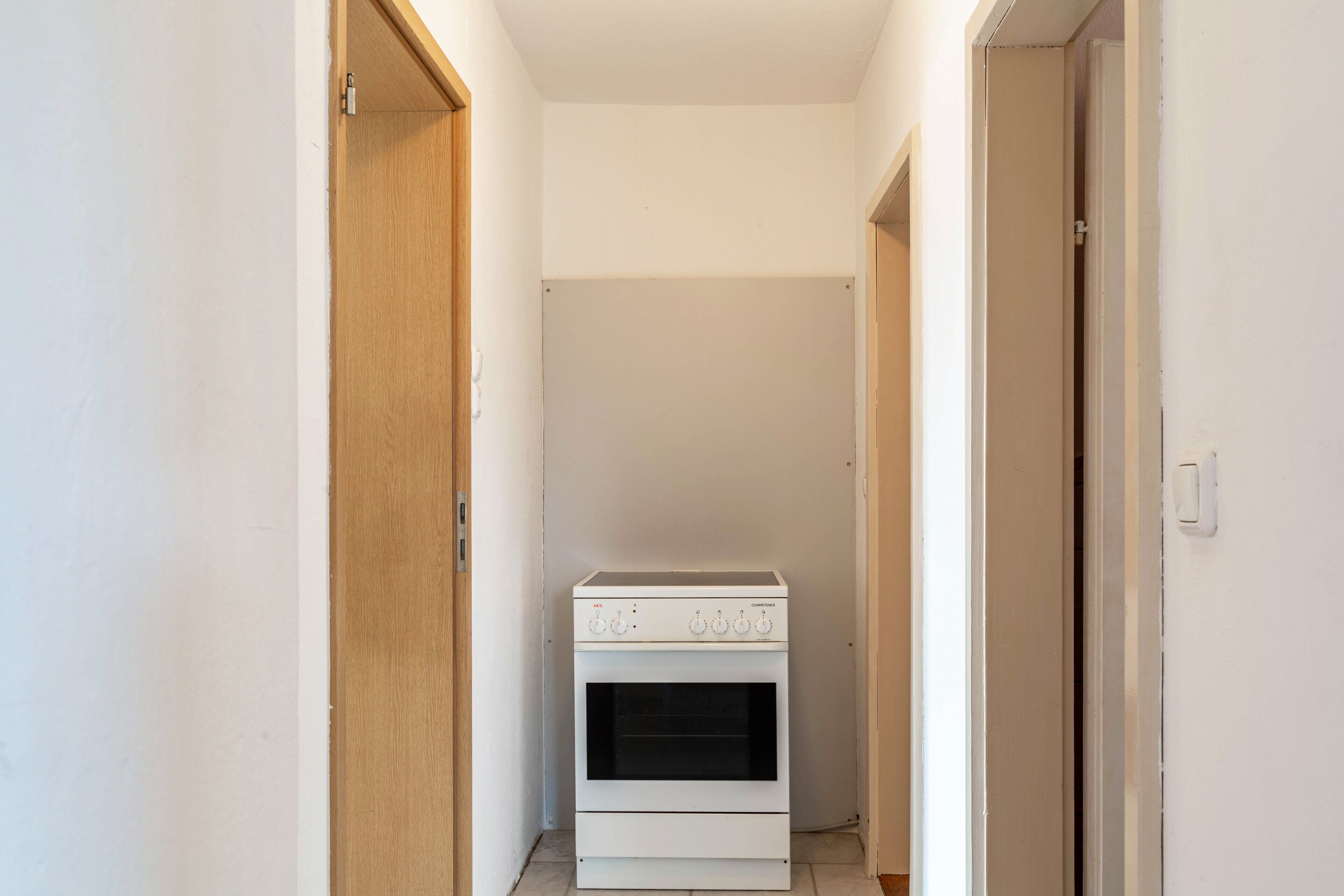 A narrow hallway with a white oven and wooden door on the left, and a white wall on the right.