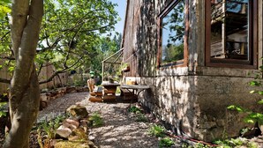 A rustic wooden building with a stone pathway, outdoor seating, and a large window.