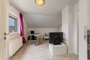 Une pièce comprenant un lit, un bureau, un ordinateur, un radiateur, une fenêtre avec des rideaux et donnant sur une maison.