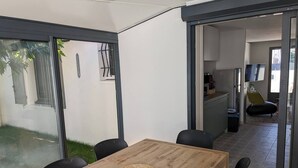 A modern dining area with a wooden table and black chairs, a woven placemat, and a glass door leading to a kitchen and living area. - Petite Bulle, a place of calm and relaxation close to the town center. (Narbonne)