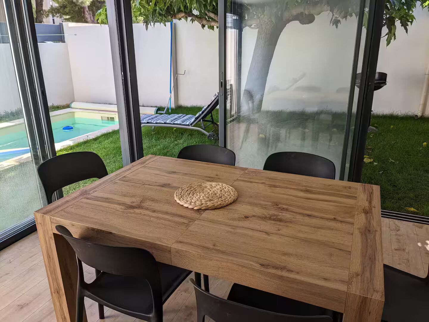 Una mesa de madera con cuatro sillas negras, un mantel tejido y una partición de vidrio con vista a una piscina y un árbol.