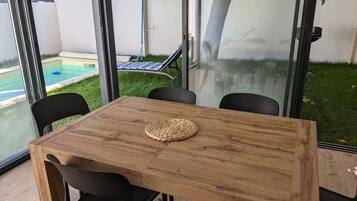 Una mesa de madera con cuatro sillas negras, un mantel tejido y una partición de vidrio con vista a una piscina y un árbol.
