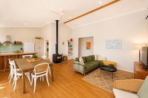 Un vaste espace de vie comprenant une table à manger en bois, des chaises blanches, un canapé vert et une cheminée noire.
