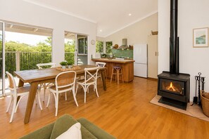 A modern kitchen with a wood-burning stove, a dining area with a table and chairs, and a view through the glass doors. - Yarrimbah Holiday Retreat  (Peppermint Grove Beach)