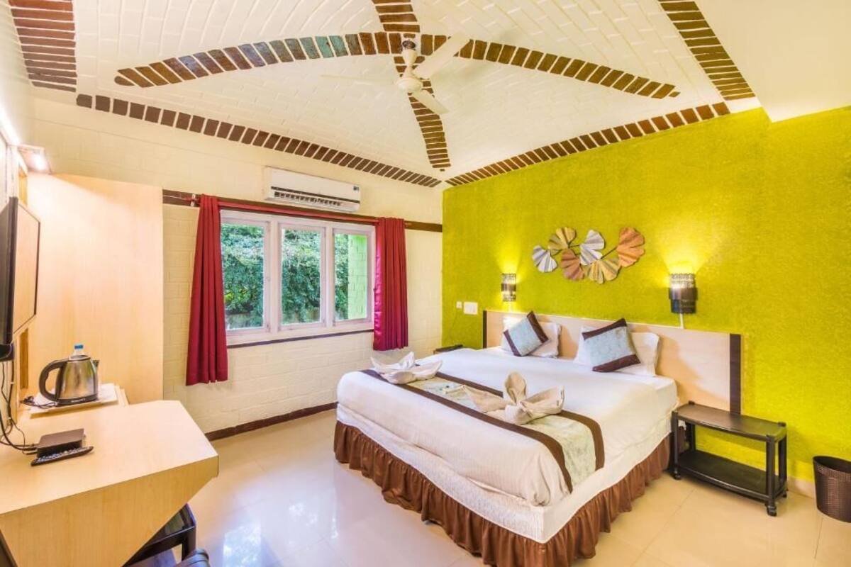 A hotel room with a large bed, a desk with a kettle, a window with curtains, and a bright yellow wall.