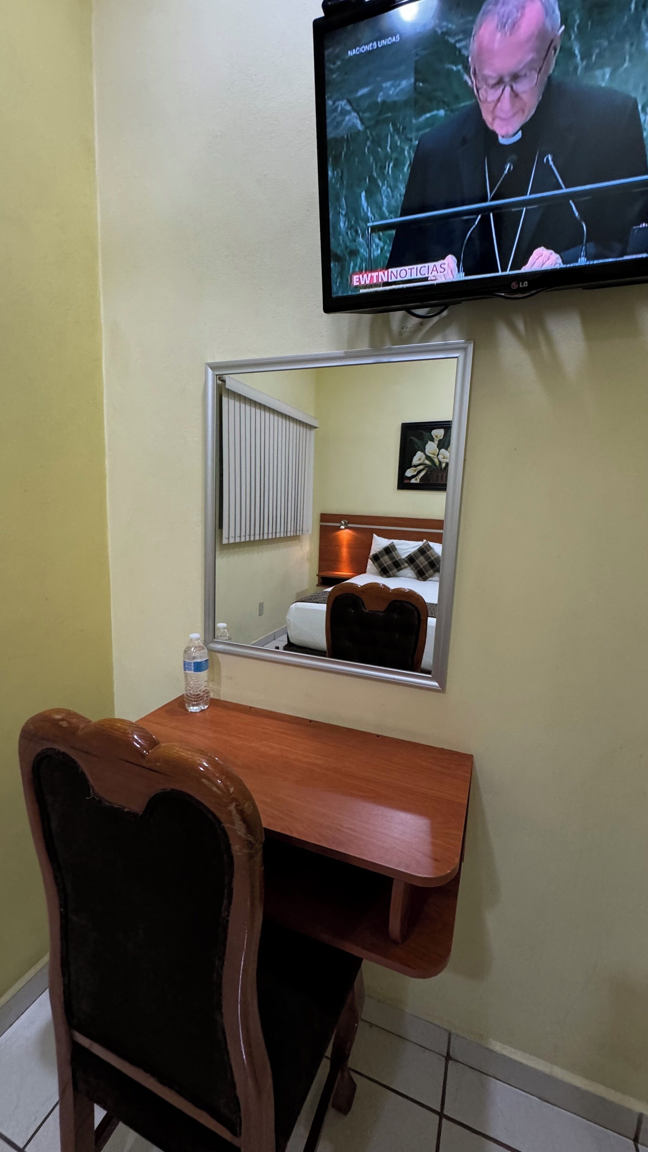 Habitación de hotel con un escritorio de madera, una silla, una cama con edredón blanco y almohadas escocesas, una mesita de noche con lámpara, y un televisor en la pared mostrando un noticiero.