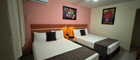 Habitación de hotel con dos camas, un ventilador de techo y dos cuadros colgados en la pared.