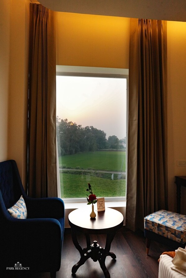 A room with a large window, a blue armchair, a small round table with a vase of flowers, and a patterned ottoman. - PARK REGENCY (Kapurthala)