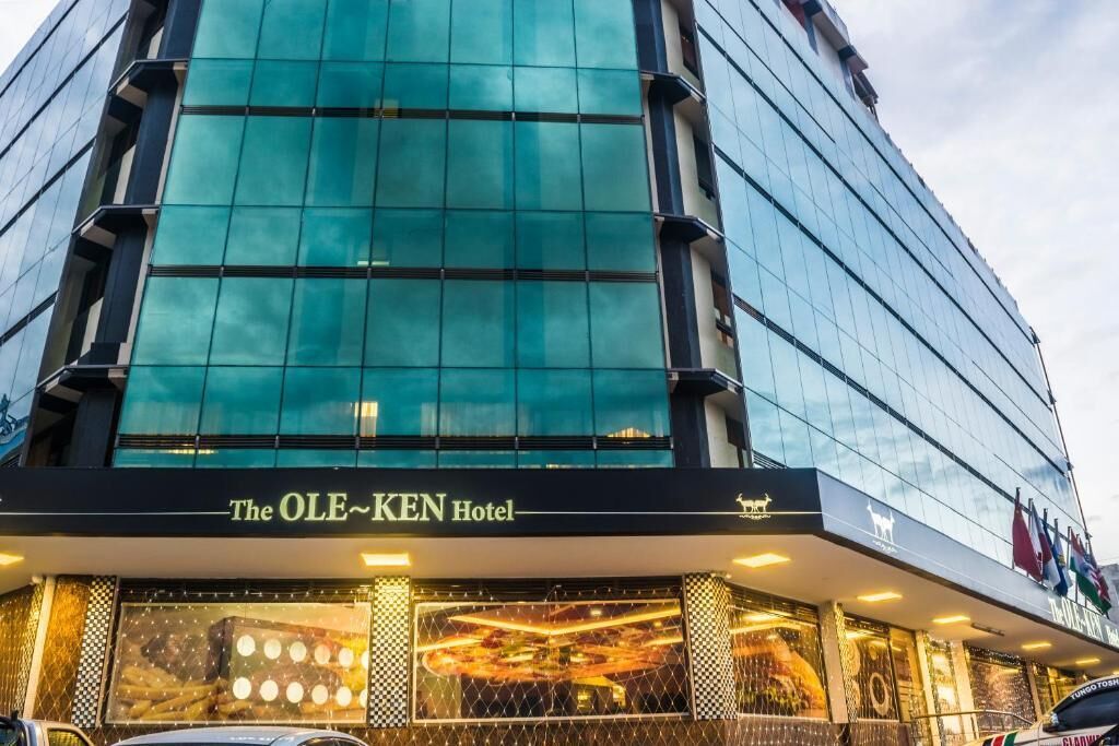 L’hôtel OLE-KEN se distingue par sa façade moderne en verre et son entrée imposante, agrémentée d’une enseigne lumineuse.