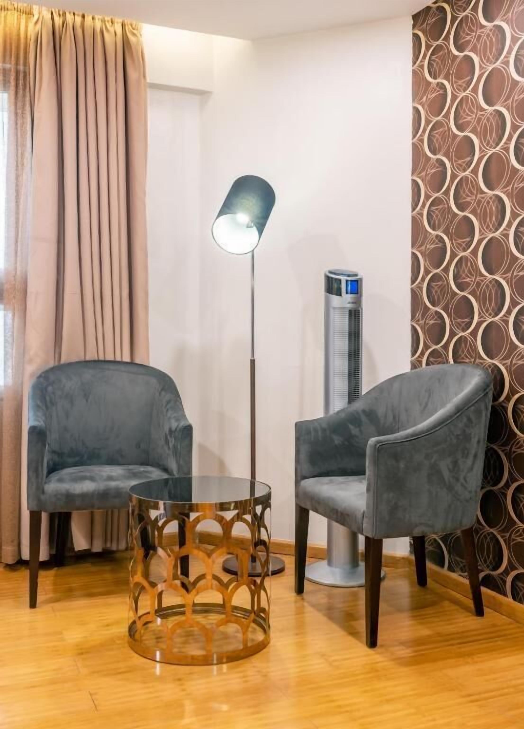 A room with two grey armchairs, a round table with a wooden base, a floor lamp, and a standing heater.