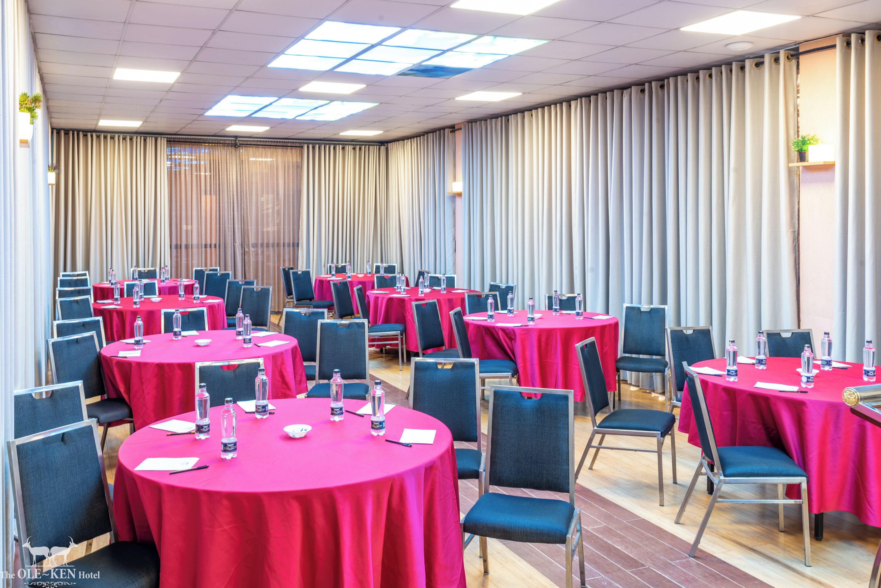 A conference room with multiple round tables covered in red tablecloths, equipped with chairs and water bottles.
