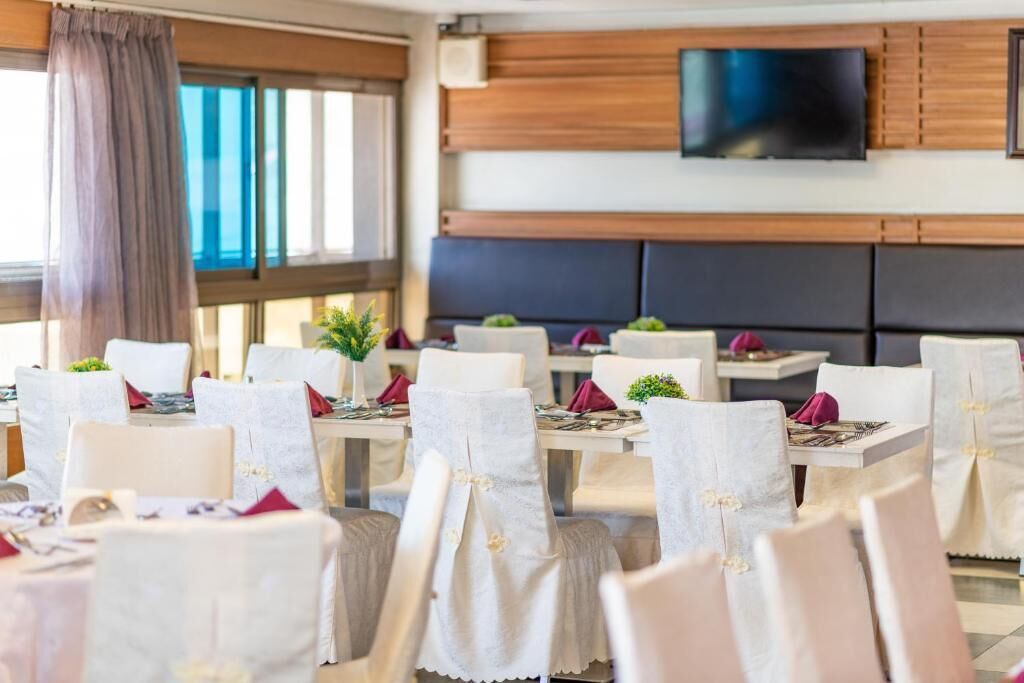 Un ristorante con sedie bianche, tavoli apparecchiati con tovaglioli rossi e un televisore montato a parete.