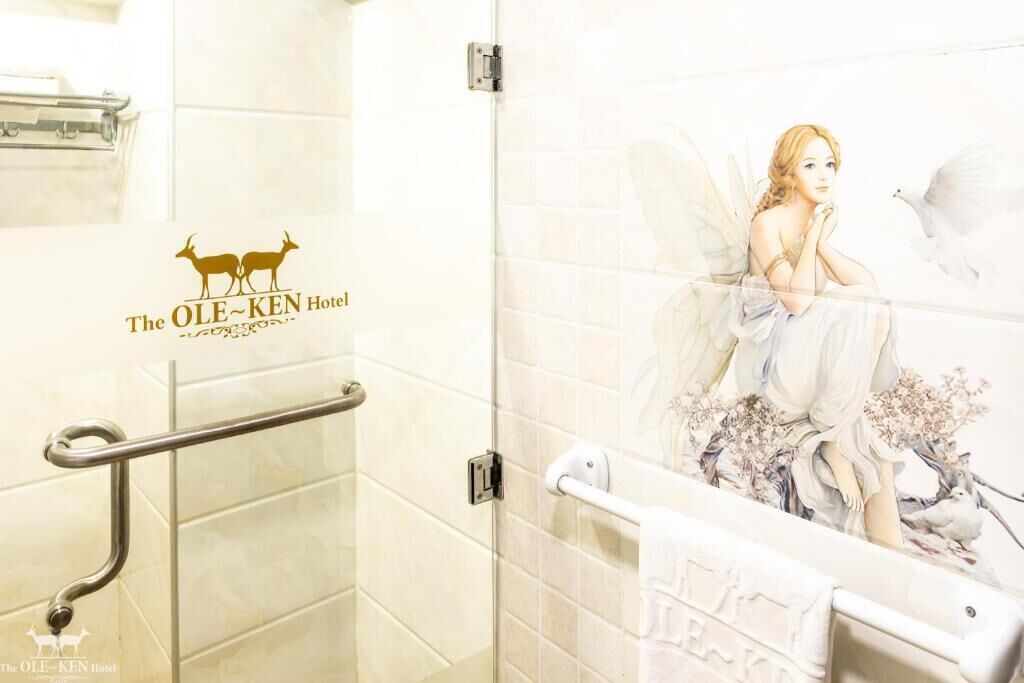 A bathroom with a towel rack, a handrail, and a mural of a woman with wings. The wall has a logo for The OLE-KEN Hotel.