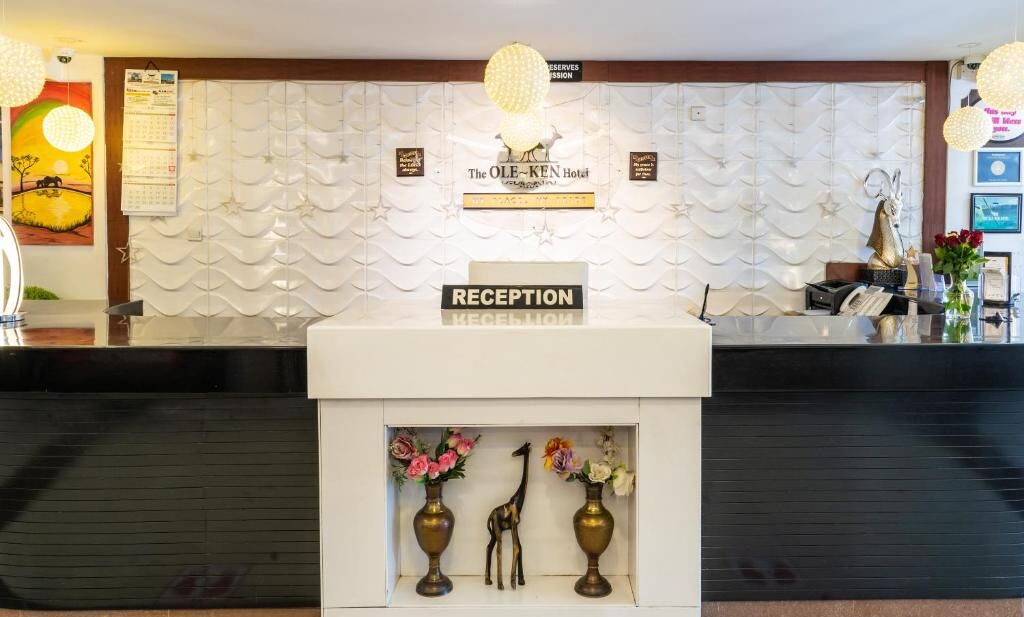 Un mostrador de recepción con un cartel que dice "RECEPTION" y "The OLE-KEN Hotel" encima. Hay jarrones decorativos con flores y una figurita de jirafa sobre el mostrador.