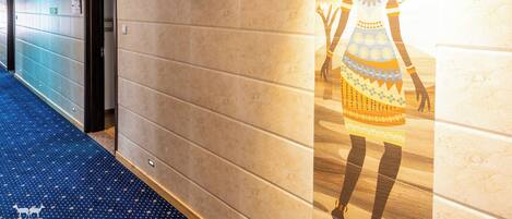 A hotel corridor with a blue carpet, a wall with a mural, and a door leading to a room.