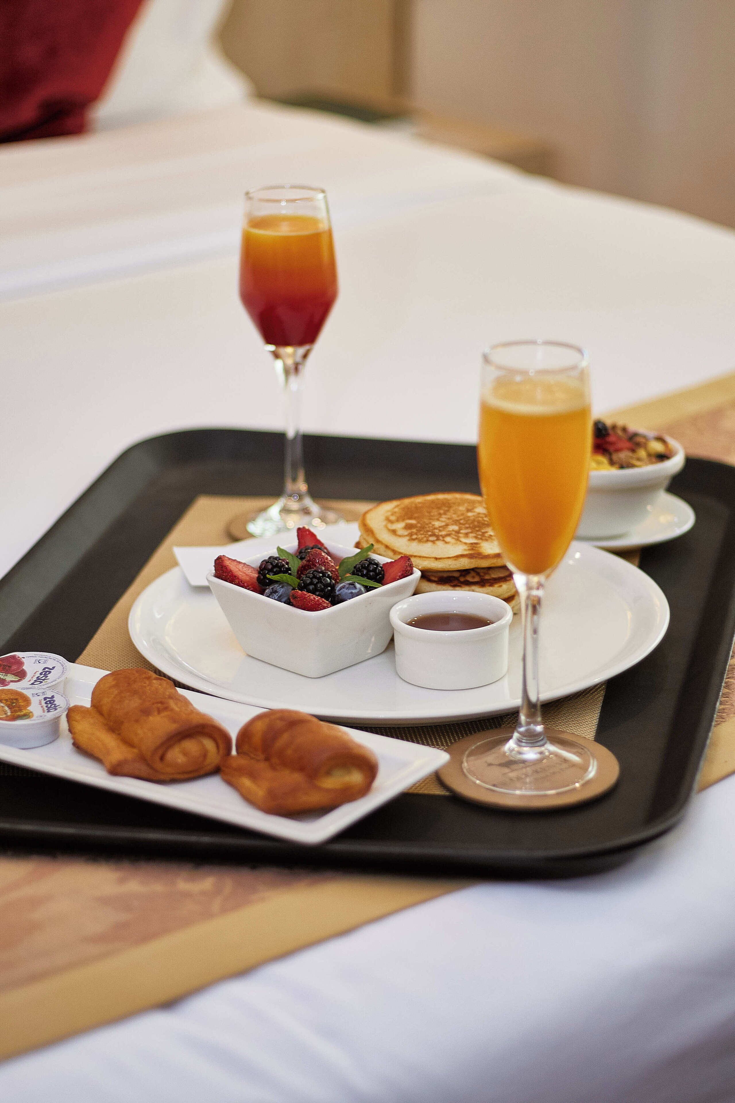 Een ontbijtkarretje met pannenkoeken, een glas sinaasappelsap, een glas champagne, een schaal gemengd fruit en croissants op een bed.