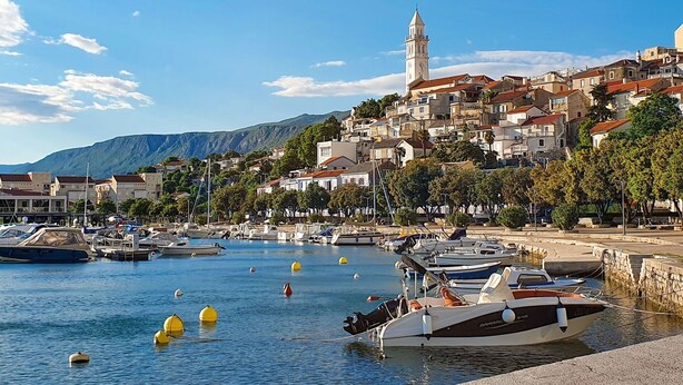 A coastal town with a prominent tower, boats docked along the waterfront, and a hilly backdrop.