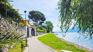 A paved walkway beside a body of water, with a stone wall and a building with arched windows on one side. - Lago Infinito by Interhome (Gravedona)